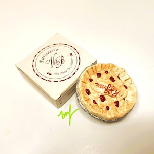 May include: A miniature cherry pie with a flaky crust and red cherries, next to a box labeled "Patisserie VdB The Cherry Pie Collection." The pie has the word "muffy" written on it. The box is white with maroon text and cherry illustrations.