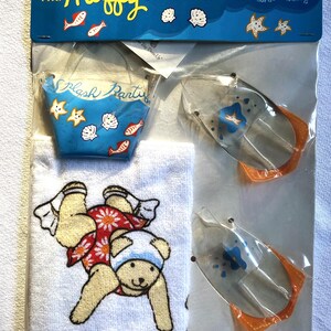 May include: A "Muffy Collection" Fun-in-the-Sun Set. The set includes a blue bucket with "Splash Party" text, a white towel with a bear design, and two clear plastic water toys with orange accents. The packaging is blue and white.