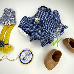 May include: A collection of Dutch-themed items, including a blue and white gingham dress, wooden clogs, a bonnet with yellow yarn braids, and a tag. The dress has white lace trim and a floral accent.