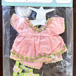 May include: A pink doll dress set from The Muffy Collection, featuring a top and shorts with floral trim. The set is packaged in a clear plastic bag with a hanger. The text "Flower Festival" is visible.