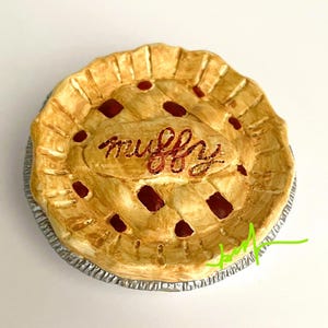 May include: A ceramic pie with a lattice crust and the word "muffy" written in red script across the top. The pie has a golden-brown crust and dark red filling, set in a silver foil pie tin.