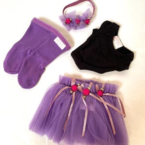 May include: A set of doll clothing including a pair of purple knit socks with a white tag labeled Happy, a black leotard with a white tag labeled Happy, a purple tulle skirt with pink ribbon and rose accents, and a matching purple tulle headband.