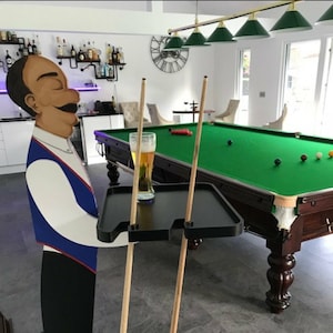 May include: A wooden figure of a waiter holding a black tray with a glass of beer on it. The figure is standing next to a pool table with two pool cues leaning against it.