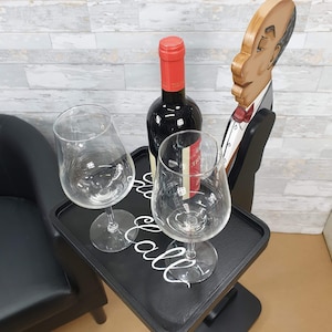May include: A black wooden serving tray with a bottle of red wine and two wine glasses. The tray has a white inscription that says "Cheers to all". A wooden figurine of a waiter is standing next to the tray.