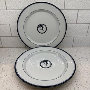 May include: Two white ceramic dinner plates featuring a dark blue rim and a thin inner blue circular line. Each plate displays a centered black circular emblem containing a stylized abstract leaf or wave design in the middle of the surface.