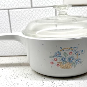 May include: White saucepan with a clear glass lid and a long white handle. The pan features a floral design with blue and red flowers in a basket. The pan is on a white countertop.