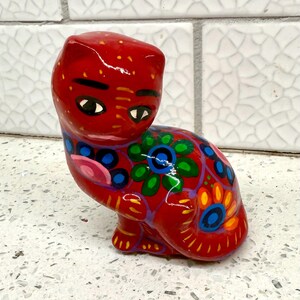 May include: A red ceramic cat figurine with a detailed floral design in blue, green, and orange. The cat has black eyes and is sitting on a white surface. The figurine is hand-painted with a glossy finish.
