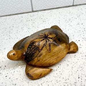 May include: Wooden turtle figurine with etched floral and geometric designs. The turtle is a light brown color with darker brown etched details. The figurine has a smooth finish and is resting on a white speckled surface.