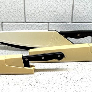 May include: Three kitchen knives with black handles and off-white sheaths. The largest knife is in a sheath, and the other two are partially sheathed. The text "THE PAMPERED CHEF" is visible on the sheaths.
