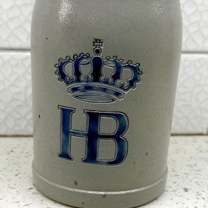 May include: A gray ceramic beer stein with a blue crown design and the letters "HB". The stein has a cylindrical shape and a slightly textured surface. The crown and letters are in a darker shade of blue.