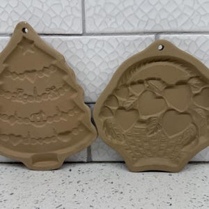 May include: Two tan ceramic cookie molds with hanging holes at the top. One mold is shaped like a Christmas tree decorated with garlands and hearts. The other mold is shaped like a basket filled with several heart-shaped fruits and leaves.