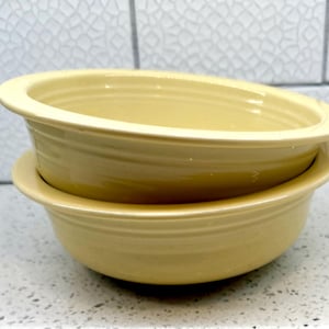 May include: Two stacked, light yellow ceramic bowls. The bowls have a rounded shape with a slightly flared rim and a ridged design around the exterior. The bowls are sitting on a speckled countertop.