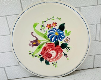 May include: A round, off-white ceramic plate with a hand-painted floral design. The flowers include a large red rose, blue and white flowers, and smaller red and yellow blossoms. A blue line borders the edge.