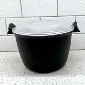May include: A black microwave rice cooker with a clear lid and black handles. The cooker is sitting on a white countertop. The lid has a white rim and the handles are on the sides of the cooker.