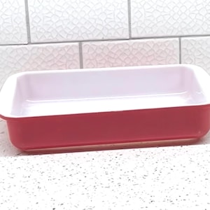 May include: A rectangular baking dish with a red exterior and a white interior. The dish is made of ceramic and is designed for oven use. The dish is approximately 9 inches long and 5 inches wide.