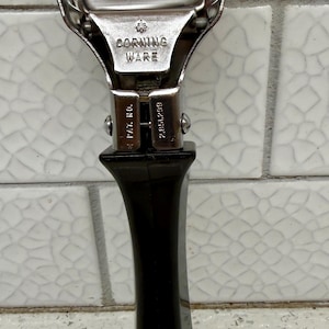 May include: A vintage Corning Ware pot handle. The handle is black and has a metal attachment with the words "CORNING WARE" stamped on it. The metal attachment has two prongs that extend upwards.