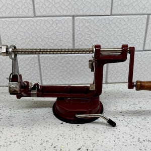 May include: A vintage apple peeler in a deep red hue, featuring a silver metal core and a wooden handle. The device has a suction base and a crank for peeling apples. The peeler is designed for kitchen use.