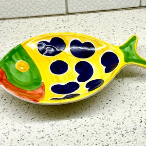 May include: A ceramic fish-shaped serving dish. The dish features a yellow base with large navy blue heart-shaped spots. The head is green with a yellow eye, and the mouth is red. The tail is green.