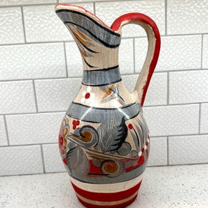 May include: A ceramic pitcher featuring a cream-colored base with hand-painted decorative patterns in red, gray, and black. The design includes floral motifs, horizontal bands, and swirling shapes. It has a curved handle and a flared pouring spout.