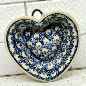 May include: Heart-shaped ceramic dish with a blue and white polka dot pattern. The rim and handle are a solid blue. The dish has a small loop for hanging. The interior is a cream color with blue and green accents.