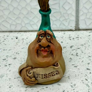 May include: A tan-colored figurine shaped like a bottle with a green neck and a brown cork stopper. The figure has a face with a large nose, red lips, and holds a scroll in its hand with the word KISSES printed in black capital letters.