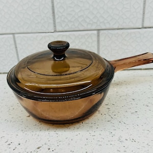 May include: A round, amber-colored glass saucepan with a matching glass lid and a single handle. The cookware features a translucent, warm brown tint and a smooth, polished finish, designed for stovetop use with a central knob on the lid.