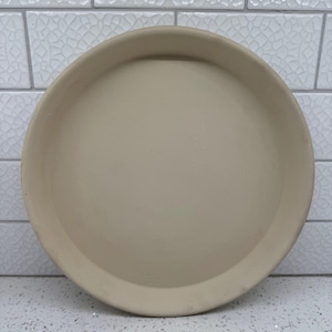 May include: A beige ceramic plate with a slightly raised edge. The plate is round and has a matte finish. The plate is sitting on a white speckled surface with a white tile background.