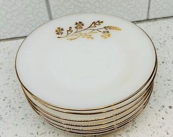 May include: A stack of white saucers with gold rims and a gold floral design. The saucers are round and appear to be made of ceramic or porcelain. The gold detailing adds a touch of elegance to the simple design.
