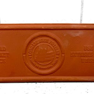 May include: A rectangular, terracotta-colored bread baking brick. The brick has rounded edges and embossed text that reads "BREAD BAKING BRICK" and "USE WITH EXTREME CARE WHEN HOT". A circular emblem is in the center.