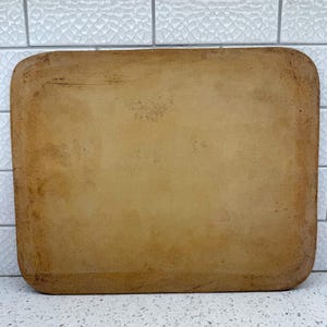 May include: A rectangular, tan-colored ceramic tray with rounded corners. The surface has a matte, earthy texture with subtle variations in tone and light brown speckling across the flat center, framed by a slightly darker, textured border.