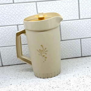 May include: A beige plastic pitcher with a handle and a yellow lid. The pitcher has a spout for pouring and a decorative floral design on the front. The pitcher is sitting on a white countertop.