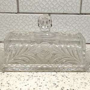 May include: A clear pressed glass butter dish with a rectangular base and a matching lid. The lid features a central circular medallion with a floral motif, flanked by leaf patterns, and topped with a faceted knob handle. The base has a diamond-cut edge.
