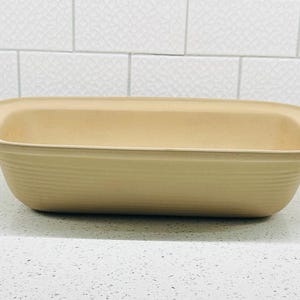 May include: A rectangular, light tan baking dish with a ribbed exterior. The dish has rounded corners and a slightly raised rim. The interior is smooth and the dish is sitting on a white surface.