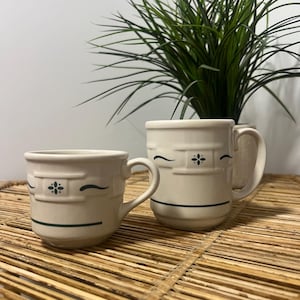 May include: Two cream-colored ceramic mugs with a glossy finish, featuring a dark green embossed geometric pattern and a horizontal line near the base. The mugs have a sturdy handle and a slightly tapered shape, showcasing a consistent design.
