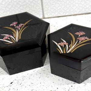 May include: Two black hexagonal lidded boxes with a glossy finish. Each box features a painted floral design on the lid, depicting pink iris flowers with long green and gold leaves. The boxes are of different sizes, with one larger than the other.