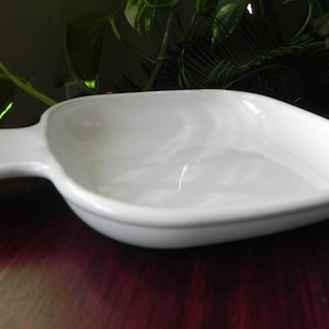 May include: A white ceramic square baking dish with rounded corners and a single integrated handle on one side. The dish features a smooth, glossy finish and a shallow basin, designed for serving or heating food items in a kitchen setting.