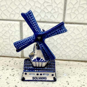 May include: A ceramic windmill figurine featuring a white base with blue painted details and a rotating blue windmill blade assembly. The front of the base displays the text SOLVANG in blue capital letters, and the structure has a glossy finish.