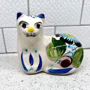 May include: A ceramic cat figurine with a white base, blue accents, and colorful floral designs. The cat has blue ears, a yellow nose, and black eyes. The figurine is hand-painted and sits on a white surface.