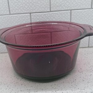 May include: A round, translucent purple glass casserole dish with integrated side handles. The bowl features a smooth, curved interior and a slightly flared rim. The glass has a deep plum hue, and the design includes molded handles on opposite sides.