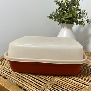 May include: A rectangular, two-tone food storage container with a lid. The base is a deep reddish-brown, and the lid is a light beige. The container is sitting on a woven bamboo tray.