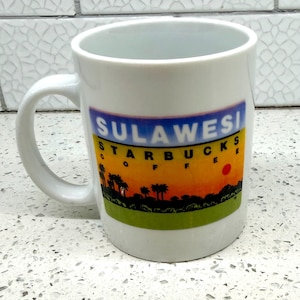 May include: White ceramic coffee mug with a handle. The mug features a colorful graphic of a sunset with palm trees and the text "SULAWESI STARBUCKS COFFEE". The mug is a souvenir.