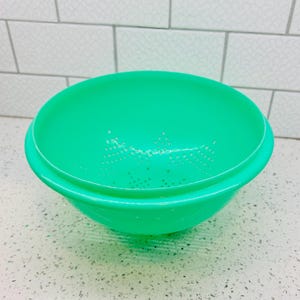 May include: A bright green plastic colander featuring a wide, circular rim and a deep, rounded basin. The interior base of the colander is perforated with a series of small, evenly spaced drainage holes arranged in a star-like pattern.