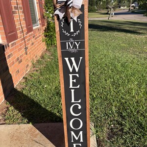 Op de afbeelding: Een hoog, rechthoekig houten veranda-bord met een frame van natuurlijk hout en een zwart middenpaneel. Het bord bevat witte tekst met "I The IVY Family WELCOME" en een lauwerkransafbeelding. Een decoratieve strik van jute en gestreept lint is bevestigd.