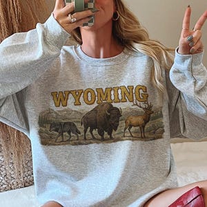 May include: Ash gray sweatshirt with the word "WYOMING" in gold lettering and a graphic of a wolf, bison, and elk. The person is wearing a white and red baseball cap and red cowboy boots.