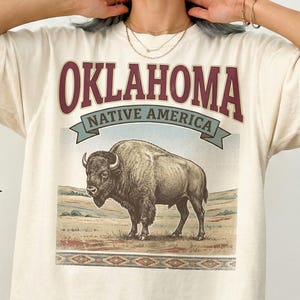 Puede incluir: Camiseta de color marfil con un diseño de bisonte en un paisaje. El texto "OKLAHOMA NATIVE AMERICA" está impreso en borgoña y verde. La camiseta tiene mangas cortas y un corte holgado.