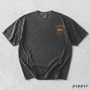 May include: Dark grey t-shirt with a crew neck and short sleeves. The shirt features a graphic on the upper chest with the word "Yellowstone" above a mountain and tree scene. The word "pepper" is written in cursive at the bottom right.