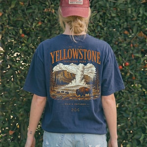May include: Navy blue t-shirt with a Yellowstone National Park graphic. The design features a bison, geyser, and forest scene. A red cap with an American flag patch is also visible.