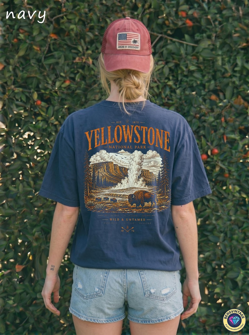 May include: Navy blue t-shirt with a Yellowstone National Park graphic. The design features a bison, geyser, and forest scene. A red cap with an American flag patch is also visible.