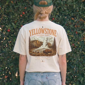 May include: Ivory-colored t-shirt with a Yellowstone National Park graphic. The design features a bison, geyser, and trees in shades of brown and orange. The shirt is short-sleeved and has the text "Yellowstone National Park" printed on the back.