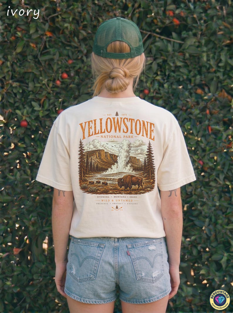 May include: Ivory-colored t-shirt with a Yellowstone National Park graphic. The design features a bison, geyser, and trees in shades of brown and orange. The shirt is short-sleeved and has the text "Yellowstone National Park" printed on the back.